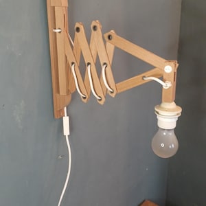 Può includere: Una lampada da parete in legno con un braccio estensibile. La lampada è in legno chiaro e ha un cavo bianco e una lampadina. È montata su un muro grigio ed è progettata per essere regolabile, con un braccio a fisarmonica.