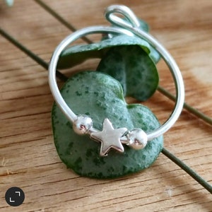 Könnte beinhalten: Ein silberner Ring mit einem Stern-Anhänger und kleinen silbernen Perlen. Der Ring liegt auf einem grünen Blatt.