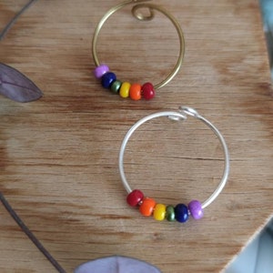 Könnte beinhalten: Zwei handgefertigte Ringe mit regenbogenfarbenen Perlen. Ein Ring ist goldfarben mit einem Spiraldesign, der andere silberfarben. Die Ringe werden auf einer Holzoberfläche mit grün-violetten Akzenten präsentiert.