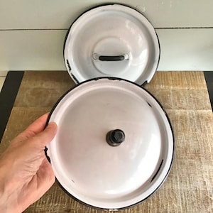 May include: Two round white enamel lids with black rims and black handles. The lids feature a smooth, glossy finish with visible signs of wear and chipping along the edges, revealing the dark metal underneath the white enamel coating.