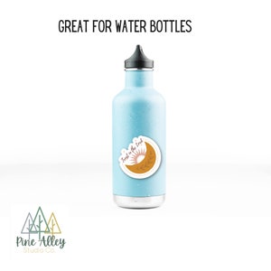 K&ouml;nnte beinhalten: Eine hellblaue Wasserflasche mit einem Aufkleber, auf dem "Trust in the good" steht, mit einem Sonnen- und Monddesign. Der Aufkleber befindet sich auf der Vorderseite der Flasche.
