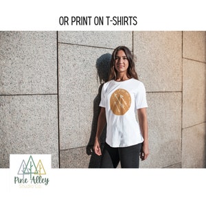 K&ouml;nnte beinhalten: Wei&szlig;es T-Shirt mit einem braunen Kreis-Motiv mit wei&szlig;en Pfeilen und dem Text "We shall meet again". Das T-Shirt wird von einer Person getragen, die vor einer Steinmauer steht. Das Bild enth&auml;lt auch ein Logo f&uuml;r Pine Alley Studio Co. mit drei stilisierten gr&uuml;nen und braunen B&auml;umen.