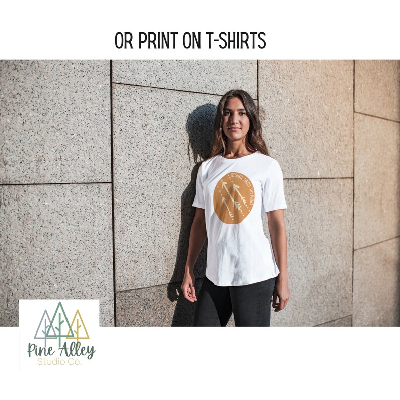 K&ouml;nnte beinhalten: Wei&szlig;es T-Shirt mit einem braunen Kreis-Motiv mit wei&szlig;en Pfeilen und dem Text "We shall meet again". Das T-Shirt wird von einer Person getragen, die vor einer Steinmauer steht. Das Bild enth&auml;lt auch ein Logo f&uuml;r Pine Alley Studio Co. mit drei stilisierten gr&uuml;nen und braunen B&auml;umen.