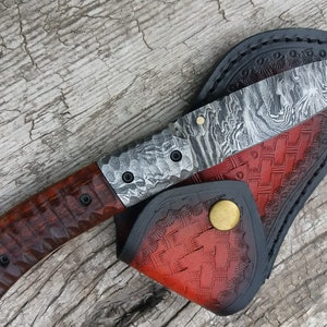May include: A handmade knife with a wood handle and a patterned steel blade. The knife is in a leather sheath with a red and black pattern.