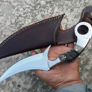 May include: A curved, silver knife with a black and white handle and a leather sheath. The knife has a ring on the handle.