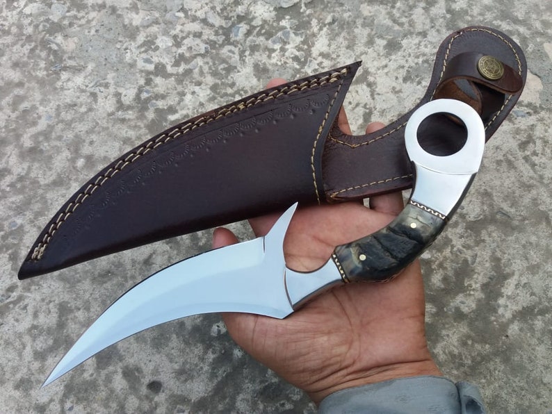 May include: A curved, silver knife with a black and white handle and a leather sheath. The knife has a ring on the handle.