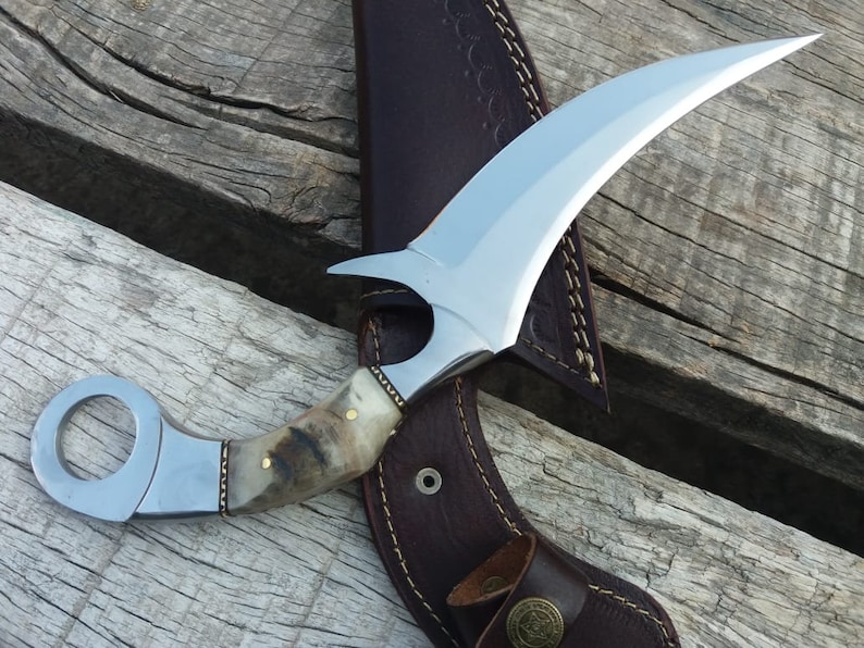 May include: A silver curved knife with a brown and white handle and a leather sheath. The knife has a ring on the handle for a finger grip.