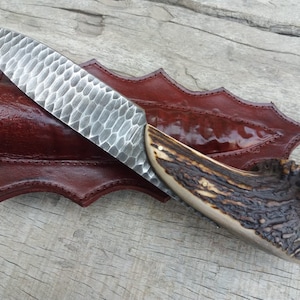 May include: A handmade knife with a hammered steel blade and a brown antler handle. The knife is in a brown leather sheath.