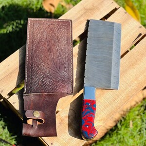 Könnte beinhalten: Ein großes, silbernes Hackmesser mit einem rot-blauen Marmor-Griff. Das Hackmesser liegt auf einer Holzoberfläche mit einer braunen Lederscheide daneben.