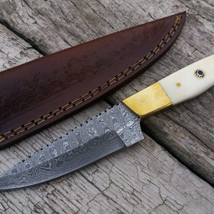 May include: A Damascus steel hunting knife with a white bone handle and brass accents. The knife comes with a brown leather sheath with decorative stitching.