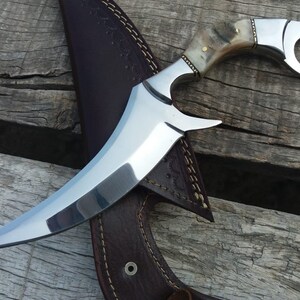 May include: A curved, silver knife with a bone handle and a ring on the top. The knife is resting on a brown leather sheath.