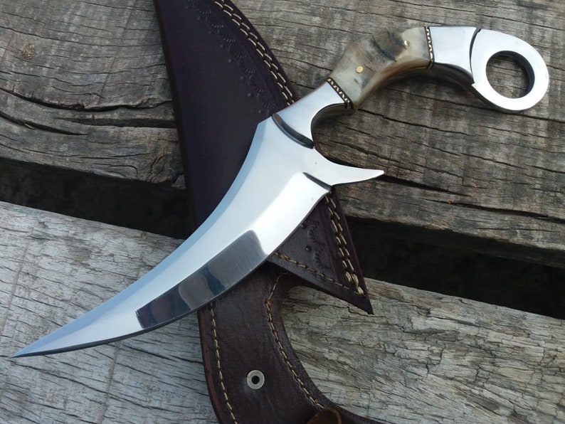 May include: A curved, silver knife with a bone handle and a ring on the top. The knife is resting on a brown leather sheath.