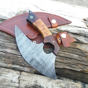 Op de afbeelding: Een handgemaakte Damascus stalen bijl met een houten handvat en een leren schede. De bijl heeft een uniek, golvend patroon op de kling.