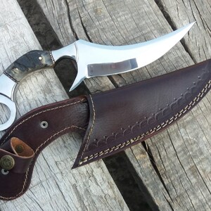 May include: A curved, silver knife with a brown and black handle and a leather sheath. The knife has a ring on the handle and the sheath has a decorative stitching pattern.