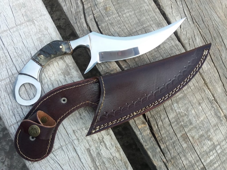May include: A curved, silver knife with a brown and black handle and a leather sheath. The knife has a ring on the handle and the sheath has a decorative stitching pattern.