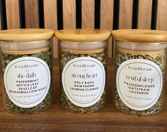 Pode incluir: Três frascos de vidro com tampas de bambu cheios de ervas secas. Cada frasco tem um rótulo branco com o texto "in a golden state" e um nome de mistura de chá diferente: "the daily", "strong heart" e "restful sleep".