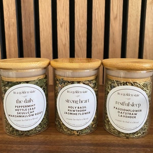 Könnte beinhalten: Drei Glasbehälter mit Bambusdeckeln, gefüllt mit getrockneten Kräutern. Jedes Gefäß hat ein weißes Etikett mit dem Text "in a golden state" und einem anderen Tee-Mischungsnamen: "the daily", "strong heart" und "restful sleep".