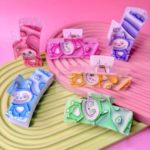 Puede incluir: Una colección de coloridas pinzas para el pelo Labubu en rosa, morado, azul, verde y amarillo. Cada pinza presenta un diseño de personaje de dibujos animados con motivos de corazón o estrella. Las pinzas son de plástico y tienen un mecanismo de resorte metálico.