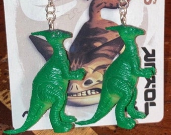 Könnte beinhalten: Ein Paar grüne Dinosaurier-Ohrringe mit silbernen Haken. Die Dinosaurier sind in entgegengesetzte Richtungen gerichtet und haben eine strukturierte Oberfläche.