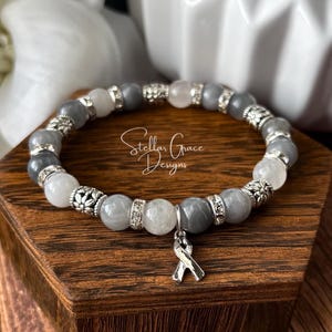 May include: A beaded bracelet featuring alternating grey and white round beads, interspersed with silver-toned metal spacers and rhinestone-encrusted rings. A silver-toned awareness ribbon charm hangs from the center. The text Stellar Grace Designs is visible.