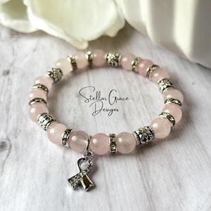 May include: A stretch bracelet featuring pale pink round beads alternating with silver-toned metal spacers and clear rhinestones. A silver-toned awareness ribbon charm with embedded rhinestones hangs from the bracelet. The text Stellar Grace Designs is visible.