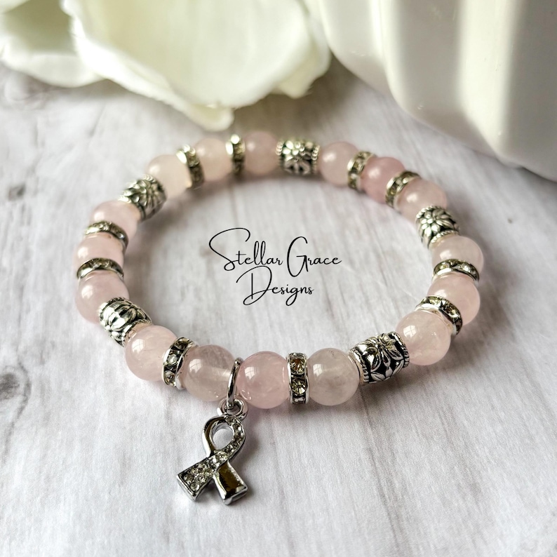 May include: A stretch bracelet featuring pale pink round beads alternating with silver-toned metal spacers and clear rhinestones. A silver-toned awareness ribbon charm with embedded rhinestones hangs from the bracelet. The text Stellar Grace Designs is visible.