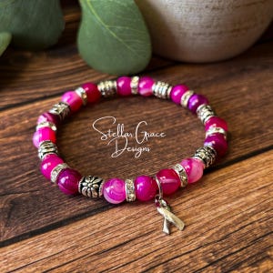 May include: A stretch bracelet featuring round, translucent pink agate beads interspersed with silver-toned metal spacers and clear rhinestone accents. A silver-toned awareness ribbon charm hangs from the bracelet, which is marked Stellar Grace Designs.