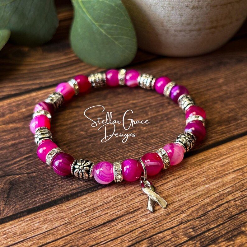 May include: A stretch bracelet featuring round, translucent pink agate beads interspersed with silver-toned metal spacers and clear rhinestone accents. A silver-toned awareness ribbon charm hangs from the bracelet, which is marked Stellar Grace Designs.