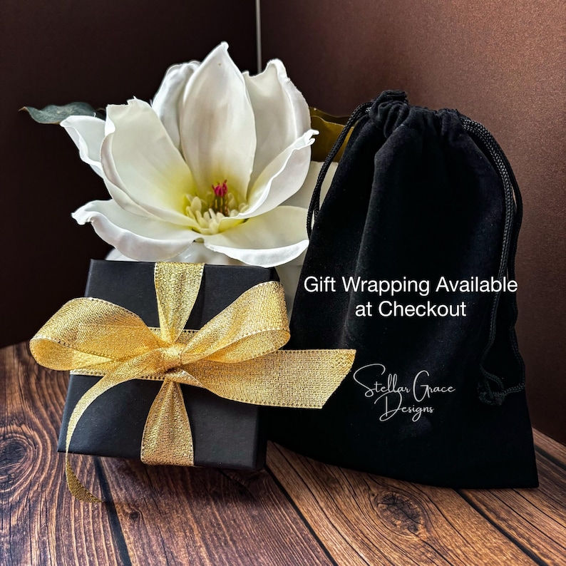 May include: A square black gift box tied with a shimmering gold ribbon, positioned next to a black velvet drawstring pouch. The pouch features the white text "Gift Wrapping Available at Checkout" and the logo "Stellar Grace Designs".