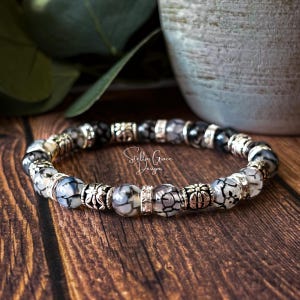 May include: A bracelet featuring a mix of black, white, and grey patterned beads, with silver-toned decorative beads. The bracelet is displayed on a wooden surface. The text "Stellar Grace Designs" is visible.