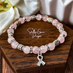 May include: A beaded bracelet featuring round, pale pink rose quartz beads alternating with silver-toned metal spacers and clear rhinestone-encrusted rings. A silver-toned awareness ribbon charm hangs from the center of the bracelet.