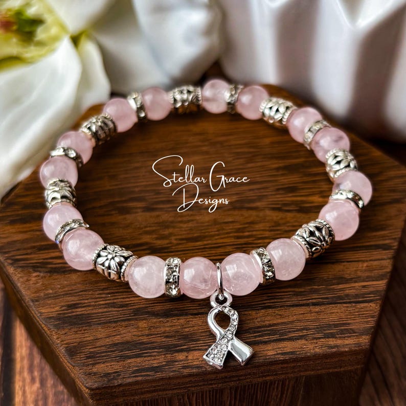 May include: A beaded bracelet featuring round, pale pink rose quartz beads alternating with silver-toned metal spacers and clear rhinestone-encrusted rings. A silver-toned awareness ribbon charm hangs from the center of the bracelet.