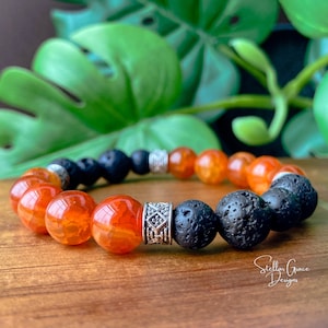 May include: A bracelet made with orange carnelian and black lava rock beads. The bracelet has a silver spacer bead with a geometric design. The bracelet is on a wooden surface.