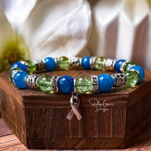 Peut inclure: Un bracelet élastique composé de perles en verre craquelé bleu et vert lime alternées, séparées par des entretoises en métal argenté et des perles décoratives à motifs floraux. Un pendentif ruban de sensibilisation argenté est suspendu.