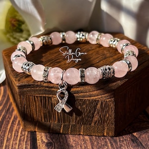 May include: A beaded bracelet featuring light pink round stones interspersed with silver-toned metal spacers and clear crystal accents. A silver-toned awareness ribbon charm encrusted with small clear crystals hangs from the center. The text Stellar Grace Designs is visible.