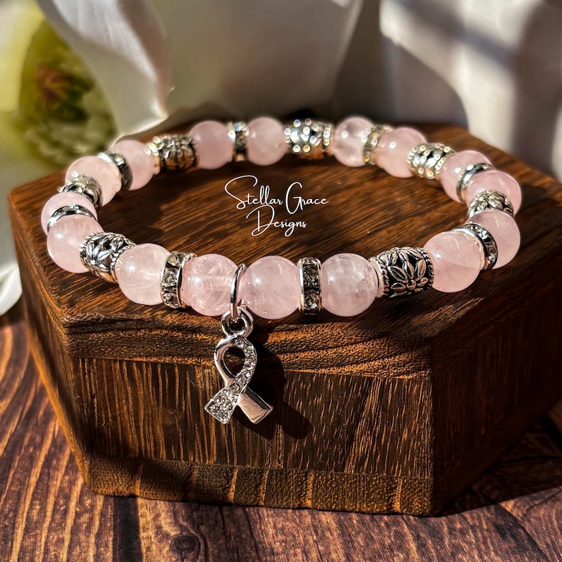 May include: A beaded bracelet featuring light pink round stones interspersed with silver-toned metal spacers and clear crystal accents. A silver-toned awareness ribbon charm encrusted with small clear crystals hangs from the center. The text Stellar Grace Designs is visible.