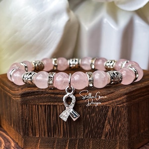 May include: A stretch bracelet featuring smooth, pale pink round beads interspersed with silver-toned metal spacers and clear crystal-encrusted rings. A silver-toned awareness ribbon charm with embedded clear crystals hangs from the center.