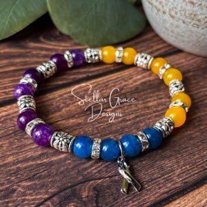 May include: A beaded bracelet featuring purple, blue, and yellow round beads separated by silver-toned metal spacers and rhinestone accents. A silver-toned awareness ribbon charm hangs from the bracelet, which is inscribed with Stellar Grace Designs.
