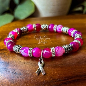 May include: A beaded bracelet featuring vibrant hot pink agate beads interspersed with silver-toned metal spacers and floral accents. A silver-toned awareness ribbon charm hangs from the center. The text Stellar Grace Designs is visible.