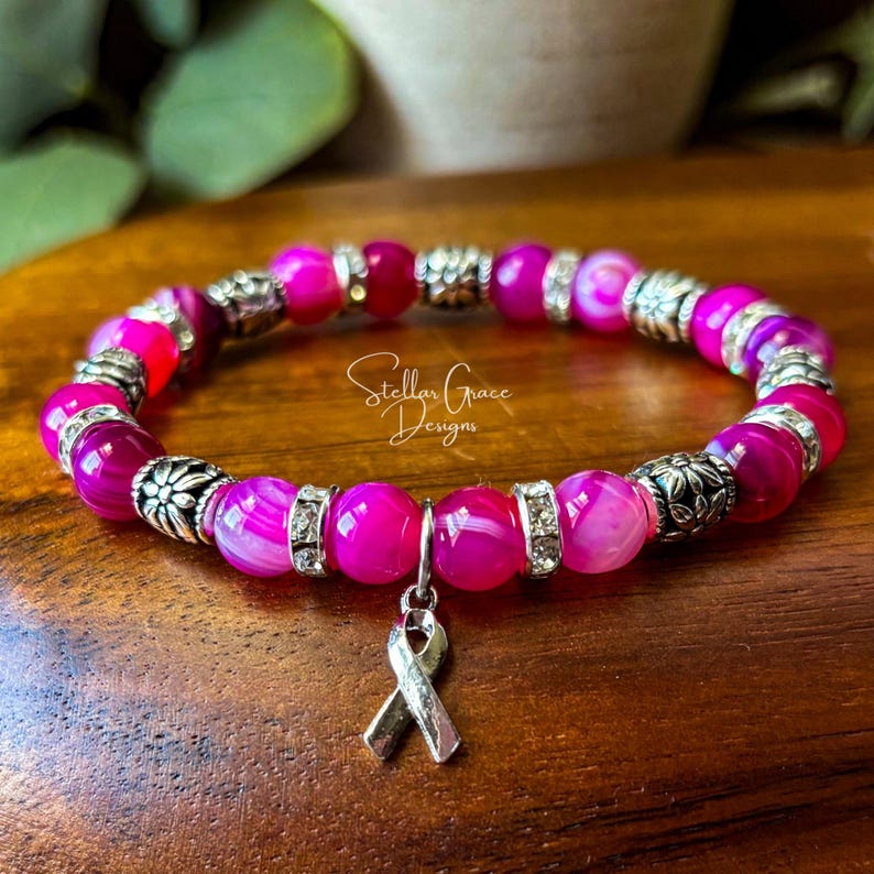May include: A beaded bracelet featuring vibrant hot pink agate beads interspersed with silver-toned metal spacers and floral accents. A silver-toned awareness ribbon charm hangs from the center. The text Stellar Grace Designs is visible.