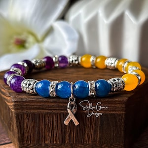 May include: A beaded stretch bracelet featuring purple, blue, and yellow round beads interspersed with silver-toned metal spacers and a silver-toned awareness ribbon charm. The bracelet is displayed on a dark wood block with the text Stellar Grace Designs.