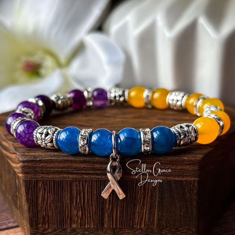 May include: A beaded stretch bracelet featuring purple, blue, and yellow round beads interspersed with silver-toned metal spacers and a silver-toned awareness ribbon charm. The bracelet is displayed on a dark wood block with the text Stellar Grace Designs.