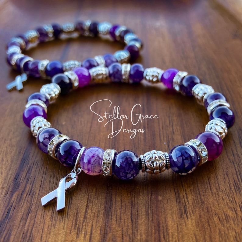 May include: Two purple agate beaded bracelets with silver accents and a silver ribbon charm. The bracelets are on a wooden surface. Stellar Grace Designs is written on the surface.