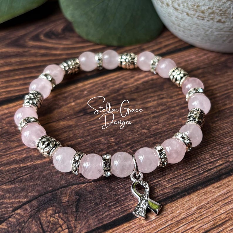 May include: A stretch bracelet featuring smooth, pale pink rose quartz beads alternating with silver-toned metal spacers and rhinestone-encrusted rings. A silver-toned awareness ribbon charm with clear rhinestones hangs from the bracelet.