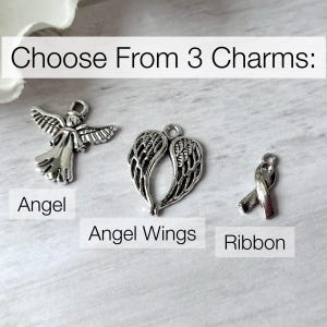 May include: Three silver-toned metal charms featuring an angel, a pair of angel wings, and a ribbon. The text "Choose From 3 Charms:" appears at the top, with labels "Angel," "Angel Wings," and "Ribbon" positioned below each respective charm.