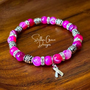 May include: A stretch bracelet featuring alternating bright pink agate beads and silver-toned metal spacers with floral detailing. A silver-toned awareness ribbon charm hangs from the bracelet, which is inscribed with the text Stellar Grace Designs.