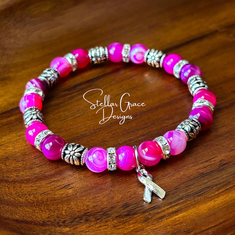 May include: A stretch bracelet featuring alternating bright pink agate beads and silver-toned metal spacers with floral detailing. A silver-toned awareness ribbon charm hangs from the bracelet, which is inscribed with the text Stellar Grace Designs.