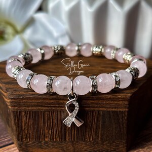 May include: A stretch bracelet featuring smooth, pale pink rose quartz beads alternating with silver-toned metal spacers and clear rhinestone-encrusted rings. A silver-toned awareness ribbon charm with clear rhinestones hangs from the center.