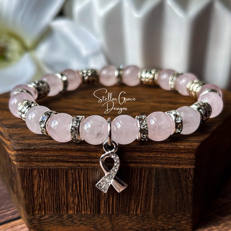 May include: A stretch bracelet featuring smooth, pale pink rose quartz beads alternating with silver-toned metal spacers and clear rhinestone-encrusted rings. A silver-toned awareness ribbon charm with clear rhinestones hangs from the center.