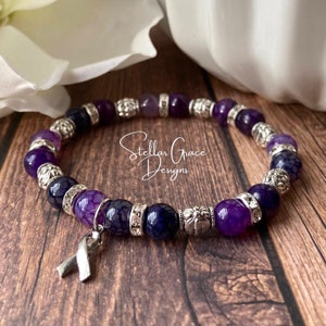 May include: Purple and silver beaded bracelet with a silver ribbon charm. The bracelet features a variety of purple agate beads and silver spacer beads. The ribbon charm is a symbol of support and awareness.